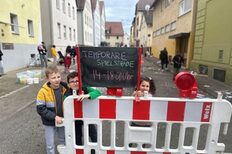 Kinder posieren hinter einer Absperrung mit einem Schild in der Hand auf dem steht: Temporäre Spielstraße 14-18 Uhr
