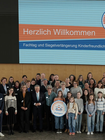 Eine Gruppe Erwachsener und Kinder steht im großen Sitzungssal im Rathaus, darunter OB Dr. Frank Nopper und die Kinderbeauftragte Maria Haller-Kindler. Kinder halten eine Attrappe des Siegels und des Stuttgarter Stadtschilds.