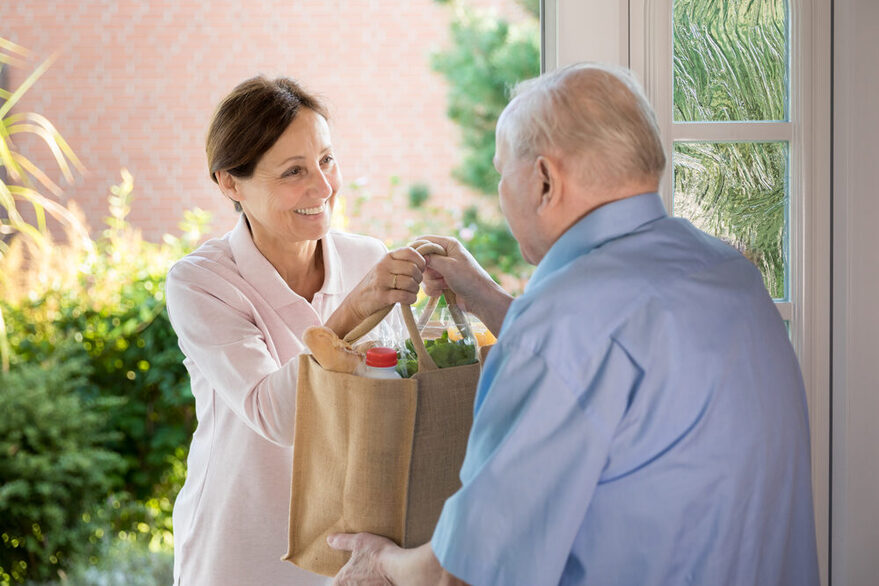 Nachbarschaftshilfe: Eine Frau überreicht einem älteren Mann eine Einkaufstasche an der Haustür.