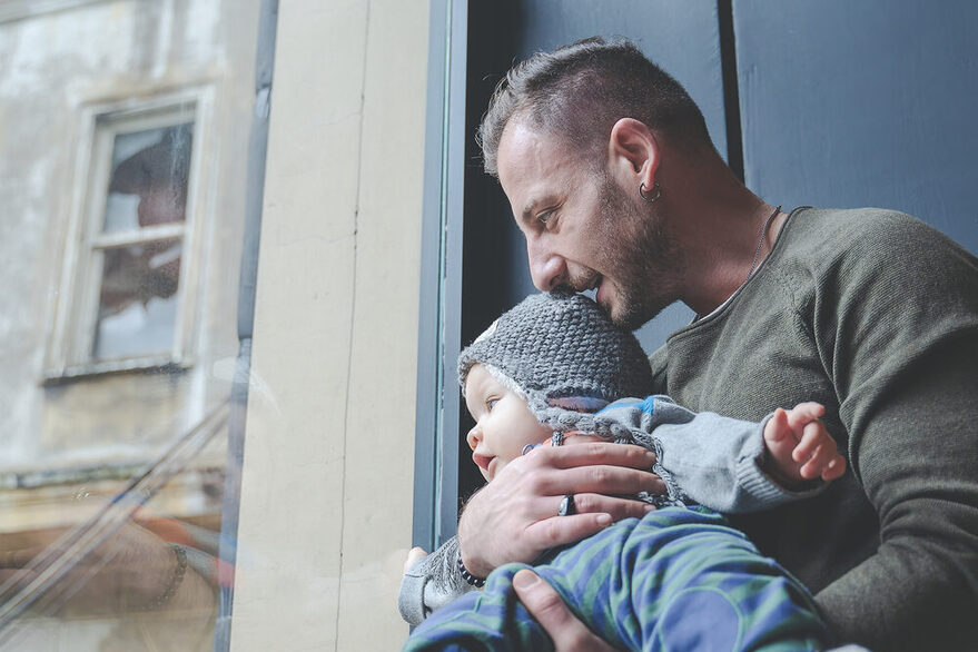 Mann hält Baby auf dem Arm und blickt aus dem Fenster.