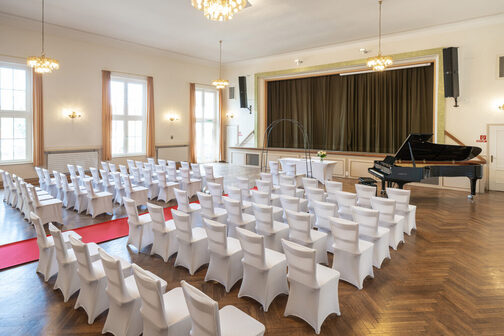 Stuhlreihen mit weißen Hussen vor einer Bühne. Im Hintergrund steht ein schwarzer Konzertflügel.