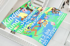 Buntes Plakat der Kinder- und Jugendbuchwochen, im Hintergrund ist das Treppenhaus der Stadtbibliothek am Mailänder Platz