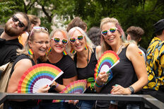 Vier Frauen mit Herzchen-Sonnenbrillen und Regenbogen-Fächern lachen in die Kamera.