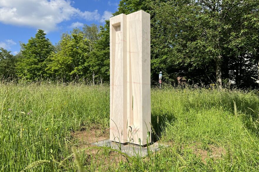 Weiße quaderförmige Marmorskulptur auf einer grünen Wiese, im Hintergrund stehen mehrere Bäume.