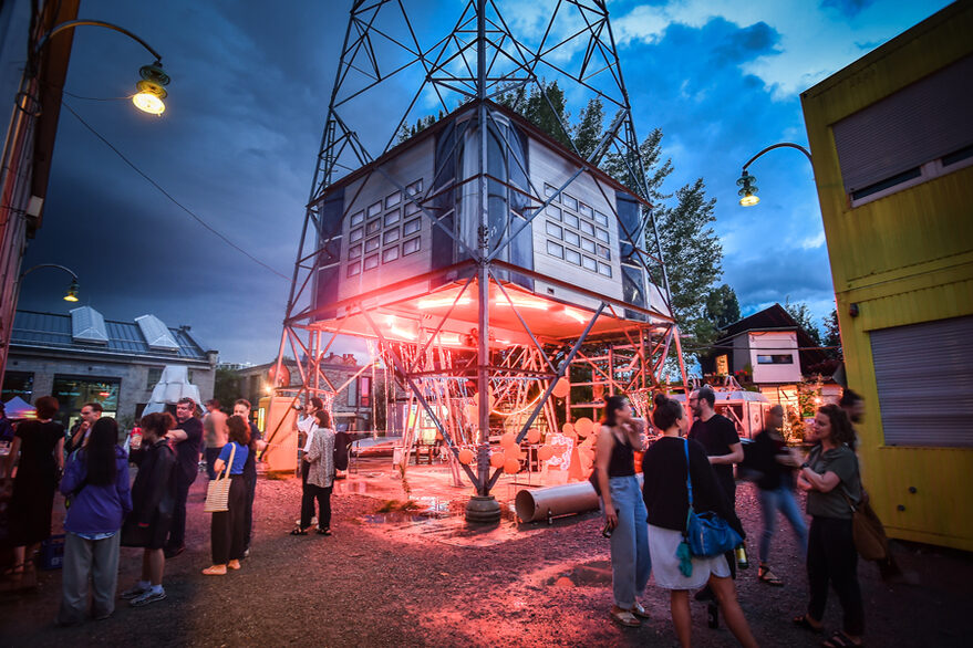 Ein rot beleutetes Kunstwerk in einem Metallturm im freien in der Dämmerung. Um Das Kunstwerk herum stehen Menschen und unterhalten sich.