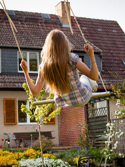 Ein Mädchen schauckelt in einem Garten hinter einem Haus.