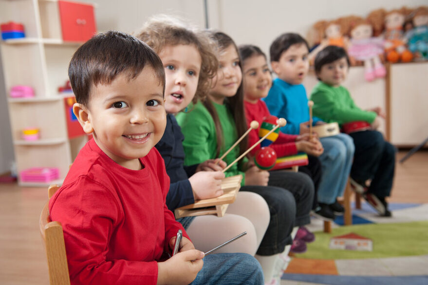 Preschool children sit in a row and hold simple instruments in their hands.