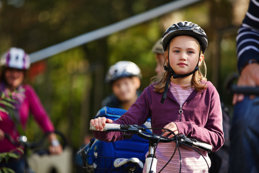 Ein Mädchen steht neben einem Fahrrad und mit Fahrradhelm auf dem Kopf vor der Kamera. Im Hintergrund sind unscharf weitere Kinder zu sehen.