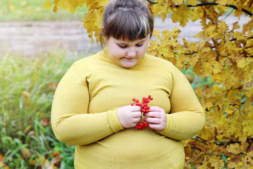 Ein übergewichtiges Mädchen steht in der Natur und hält einige Beeren in der Hand. Das Mädchen hat einen traurigen Gesichtsausdruck.
