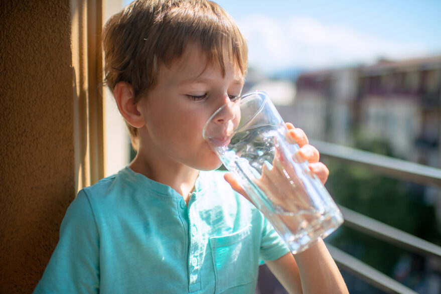 Ein kleiner Junge steht auf dem Balkon und trinkt ein Glass Wasser.