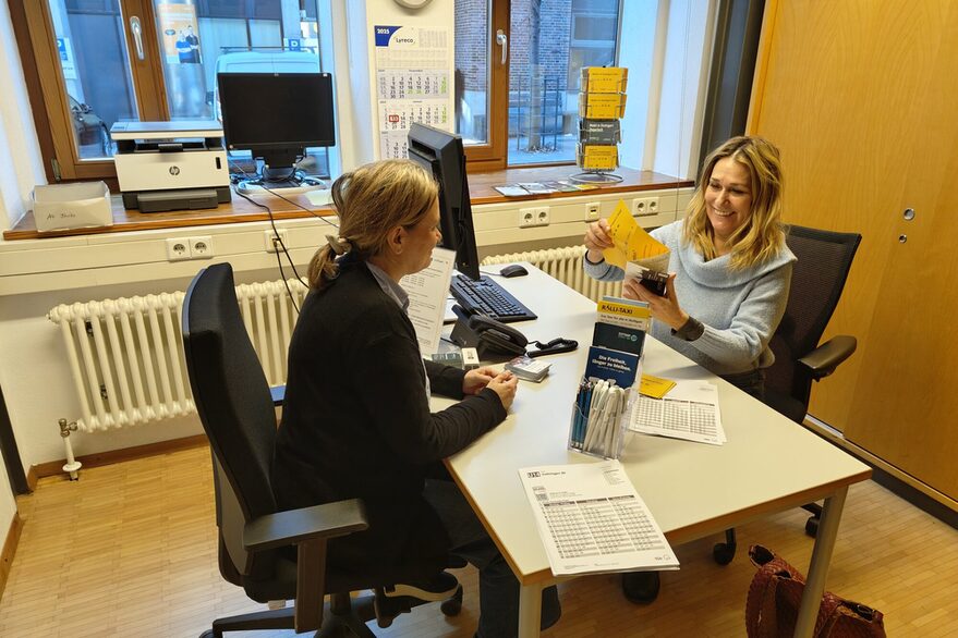 Zwei Frauen sitzen an einem Tisch. Zwischen ihnen liegen mehrere Fahrplanaushänge auf dem Tisch, ebenso wie mehrere Flyer. Die rechts sitzende Frau zeigt der anderen etwas in einem Flyer.