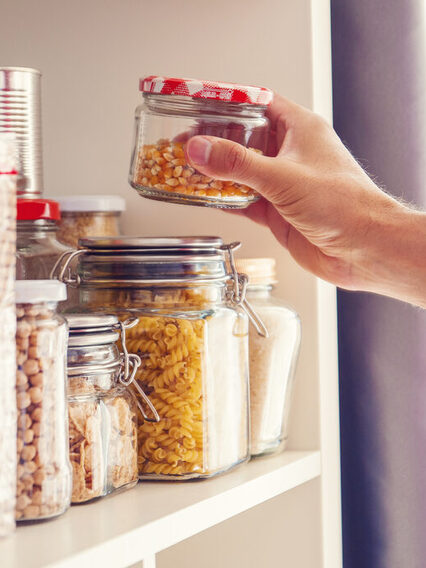 Vorratskammer: Eine Hand greift in ein Regal mit Gläsern, in denen sich Nudeln, Nüsse und andere Lebensmittel befinden.