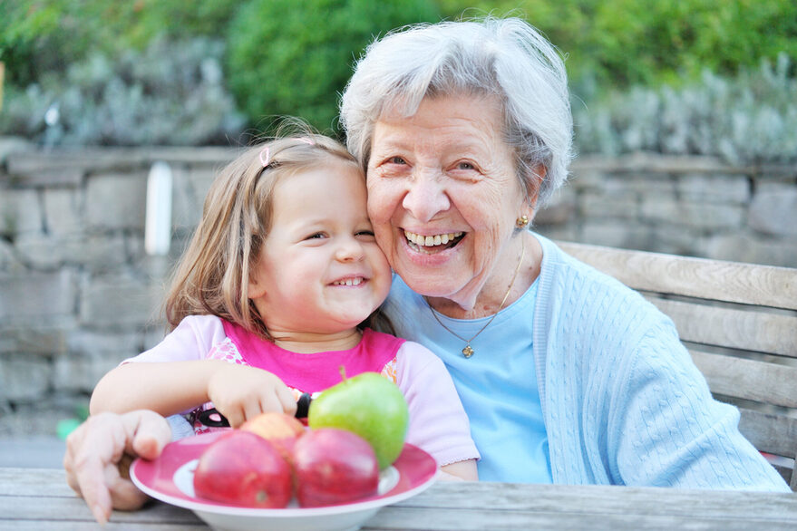 Seniorin hat Enkelkind im Arm, beide sind fröhlich, ein Teller mit Äpfel steht auf dem Tisch