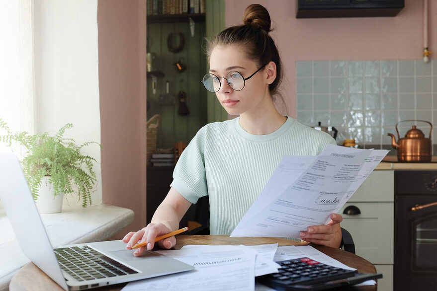 Junge Frau mit Brille sitzt am Tisch vor ihrem Laptop und hält einen Zettel in der Hand.