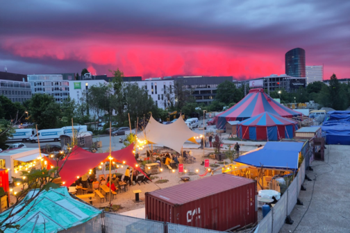 Bunte Zeltdächer und ein Zirkuszelt auf einem Platz, umgeben von weißen und grauen Hochhäusern, vereinzelt stehen Container und Transporter. Pinker Abendhimmel im Hintergrund.