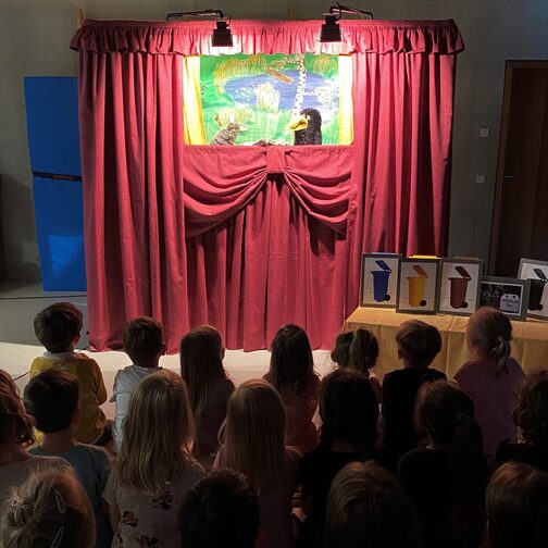 Eine Puppentheaterbühne mit rotem Vorhang. Kinder schauen beim Theaterstück mit zwei Handpuppen, einem Hasen und Raben, zu.