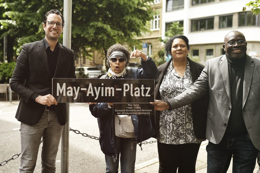 Zwei Männer (einer hiervon der Erste Bürgermeister Dr. Mayer) und zwei Frauen halten ein großes Straßenschild hoch mit dem Namen May-Ayim-Platz.
