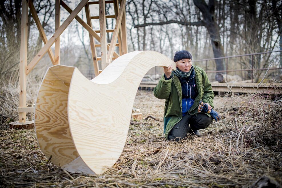 Porträt der Künstlerin Anne Duk Hee Jordan: Sie kniet im Wald mit einer großen Holzskulptur am Ohr.