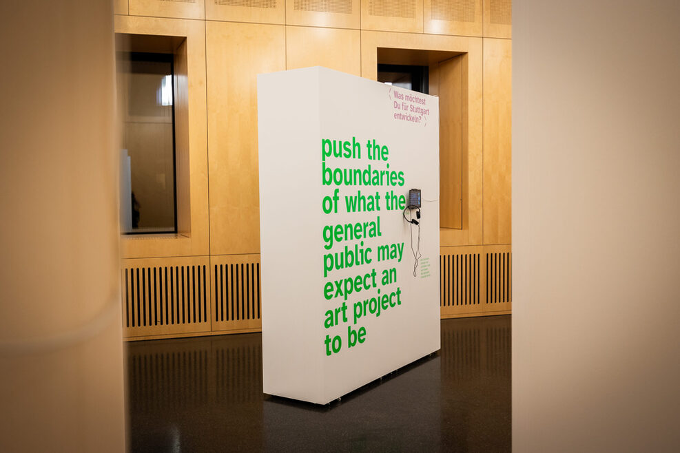 Eine weiße Wand in einem mit Holz verkleideten Ausstellungsraum auf der mit grüner Farbe "push the boundaries of what the general public may expect an art project to be" steht.