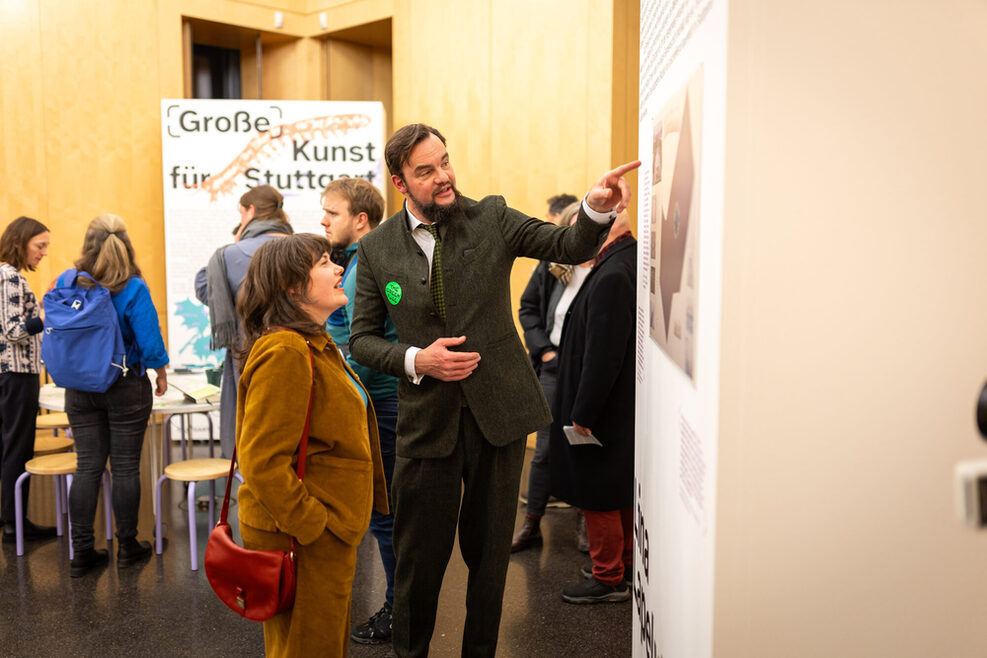 Kulturamtsleiter Marc Gegenfurtner im Gespräch mit Ruth Ewan. Herr Gegenfurtner deutet mit dem Finger auf ein Bild auf einer großen Stellwand.