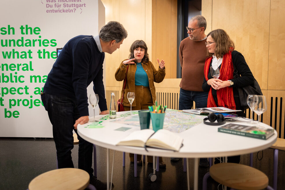 Ruth Ewan im Gespräch mit drei Besucher*innen der Ausstellung im StadtPalais. Die Personen stehen um einen runden, weißen Tisch, auf dem Gläser eine Karte, Bücher und weitere Gegenstände liegen.