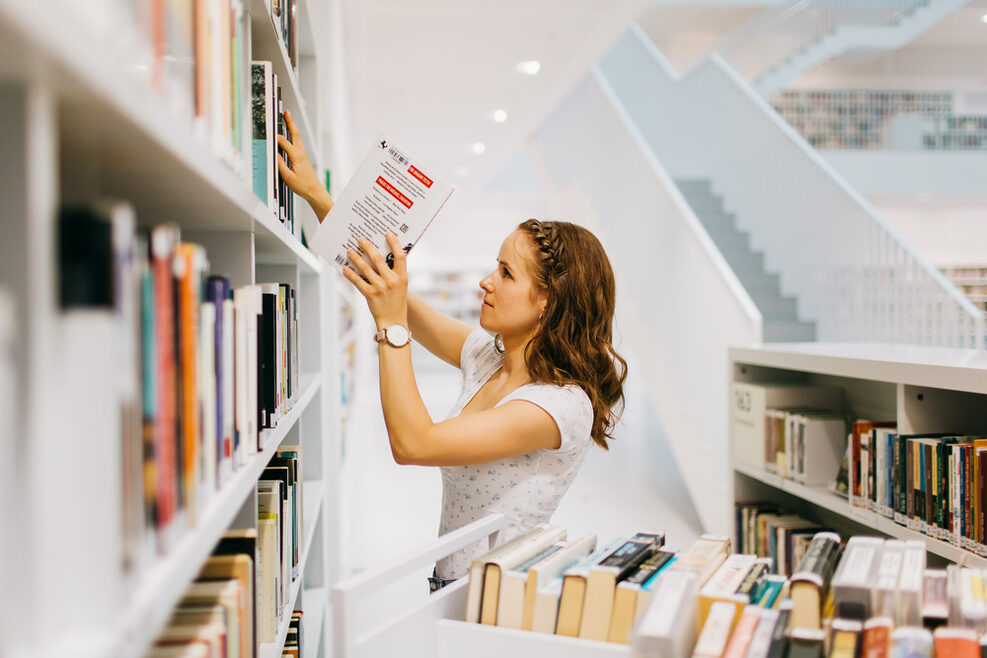 Eine Person stellt Bücher in ein Bibliotheksregal. Vor ihr steht ein Bücherwagen auf dem weitere Bücher stehen.