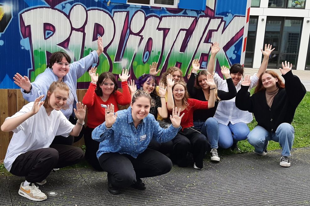 Mehrere Jugendliche in der Hocke mit erhobenen Händen vor einem Bauwagen mit einem Graffiti "Bibliothek".