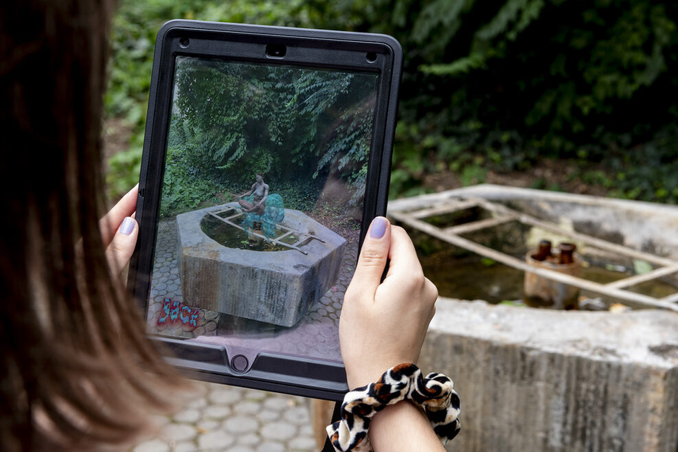 Zwei Hände halten ein Tablet vor einen Brunnen. Der Bildschirm des Tablets zeigt eine Animation von einer Figur die auf dem Brunnen sitzt.