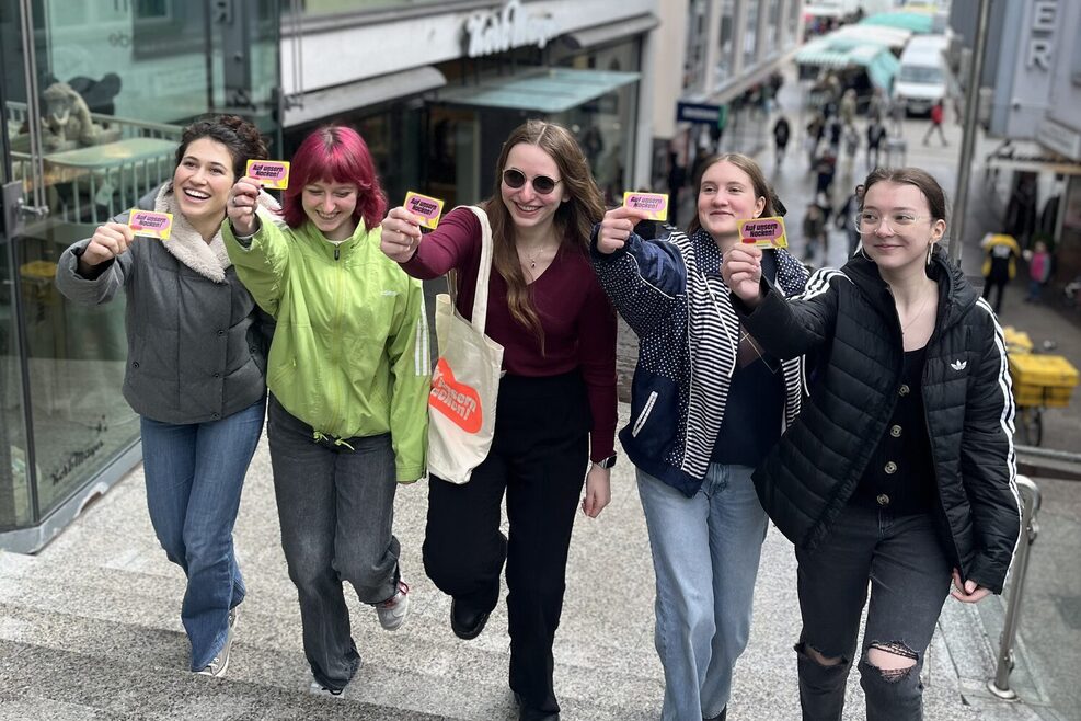 Foto: Fünf Jugendliche laufen nebeneinander eine Treppe nach oben auf die Kamera zu. Sie lachen und halten Kulturpass-Karten in die Kamera. Rechts und links ist die Treppe von Häuserfronten flankiert.