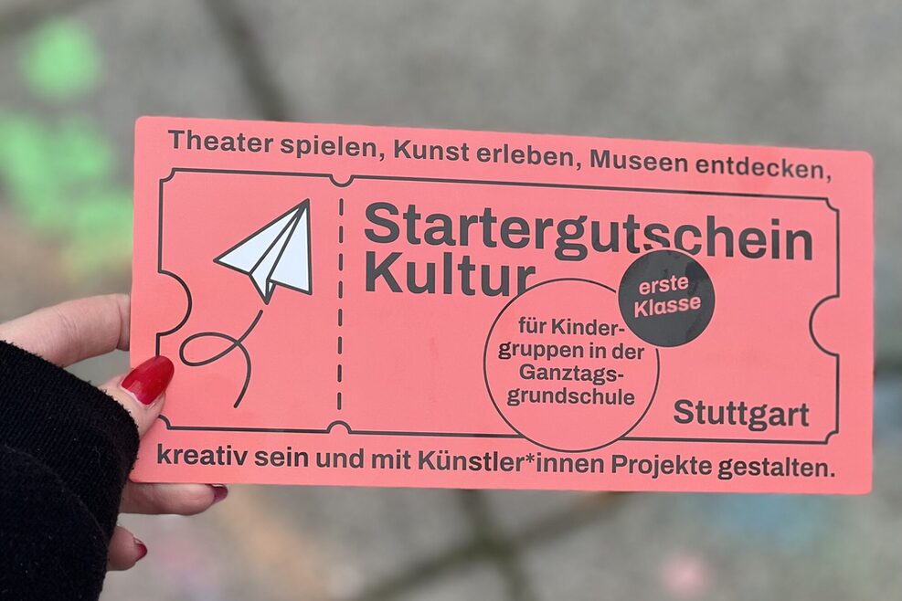 Foto: Eine Hand hält die Werbepostkarte für den Startergutschein Kultur. Auf der Karte steht: Startergutschein Kultur, „erste Klasse“ und „für Kindergruppen in der Ganztagsgrundschule“ und Theater spielen, Kunst erleben, Museen entdecken - kreativ sein und mit Künstler*innen Projekte gestalten.