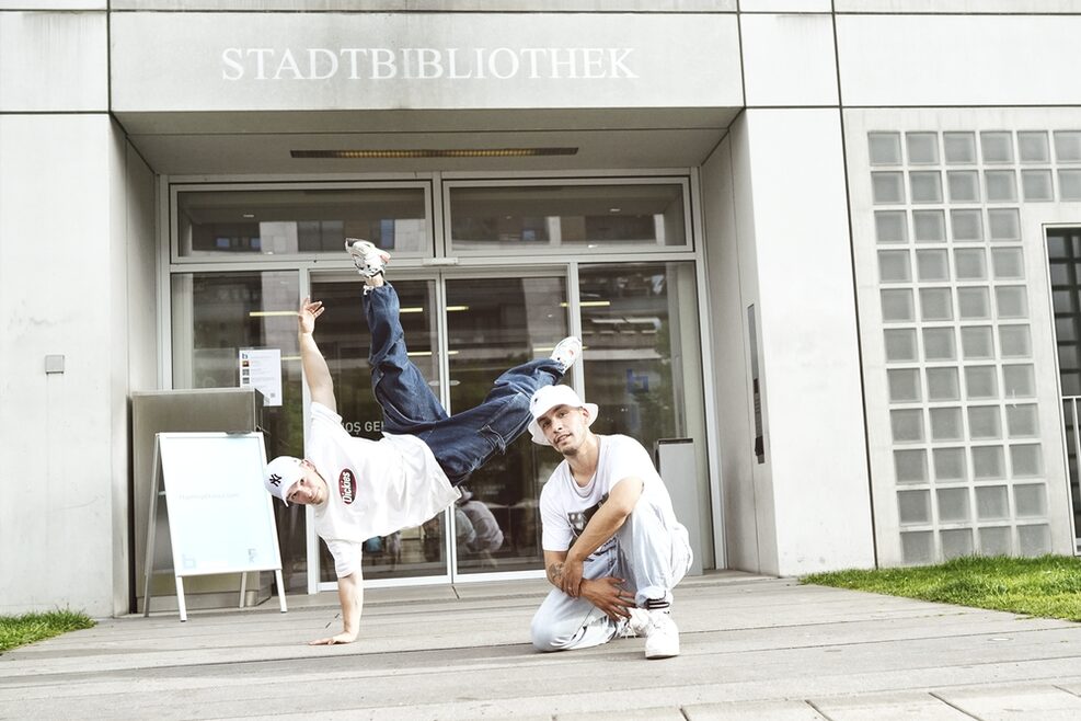 Zwei Personen vor der Stadtbibliothek Stuttgart, eine Person macht ein Kunststück auf einer Hand, die andere Person kniet auf einem Bein davor.