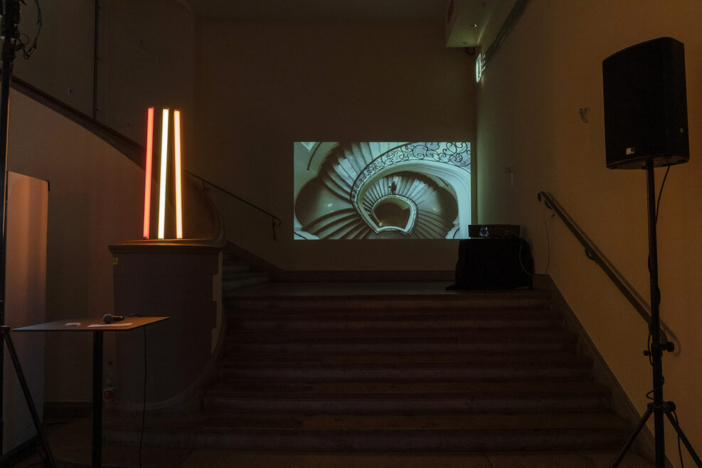 Licht- und Videoinstallation. Auf einer Leinwand in einem dunklen Raum ist ein Treppenhaus von oben zu sehen.