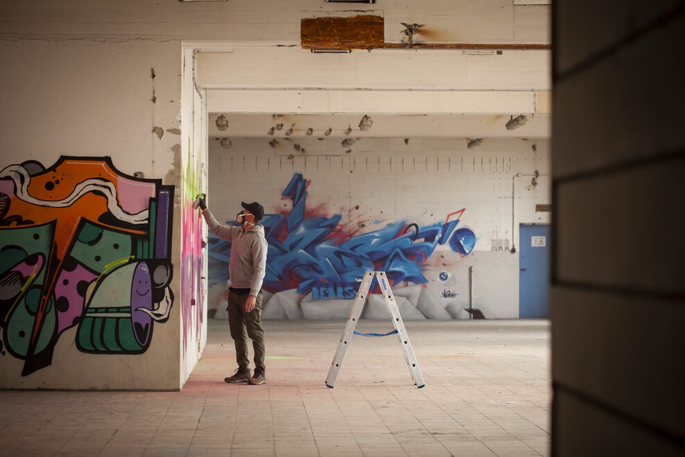 Ein Sprayer bei der Arbeit in einem alten Fabrikgebäude, hinter ihm steht eine Trittleiter.