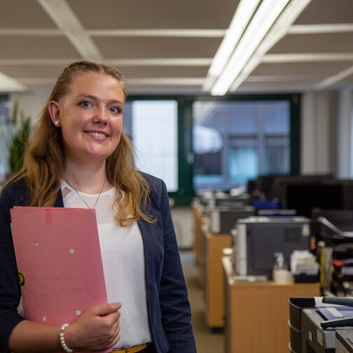 Larissa steht in einem Büro und hält eine Umlaufmappe in der Hand. Sie lächelt in die Kamera.