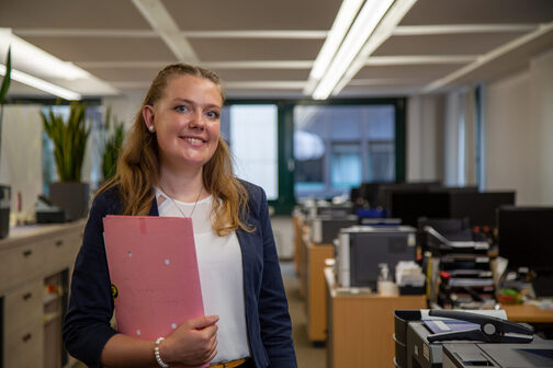 Larissa steht in einem Büro und hält eine Umlaufmappe in der Hand. Sie lächelt in die Kamera.