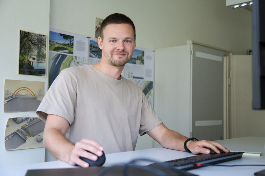 Stadtplaner Yannik sitzt mit kurzen Haaren und beigem Shirt in einem Büro, während er einen PC bedient und freundlich in die Kamera schaut.