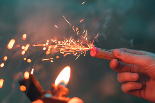 Eine Hand hält einen brennenden Böller, davor ein brennendes Feuerzeug.