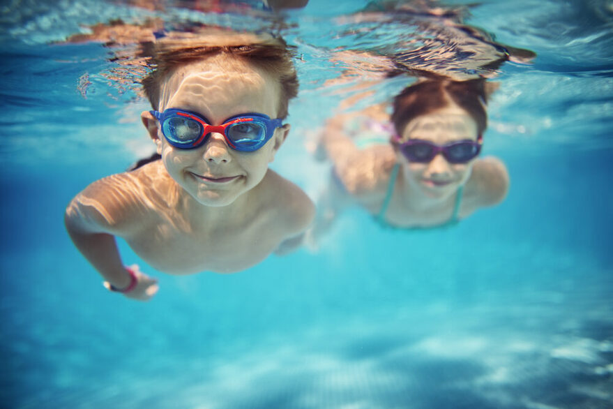 Zwei Kinder tauchen mit Schwimmbrillen unter Wasser und schwimmen lächelnd auf die Kamera zu.