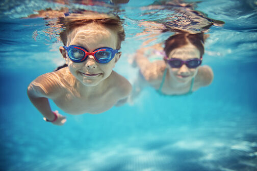 Two children with swimming goggles dive underwater and swim towards the camera, smiling.