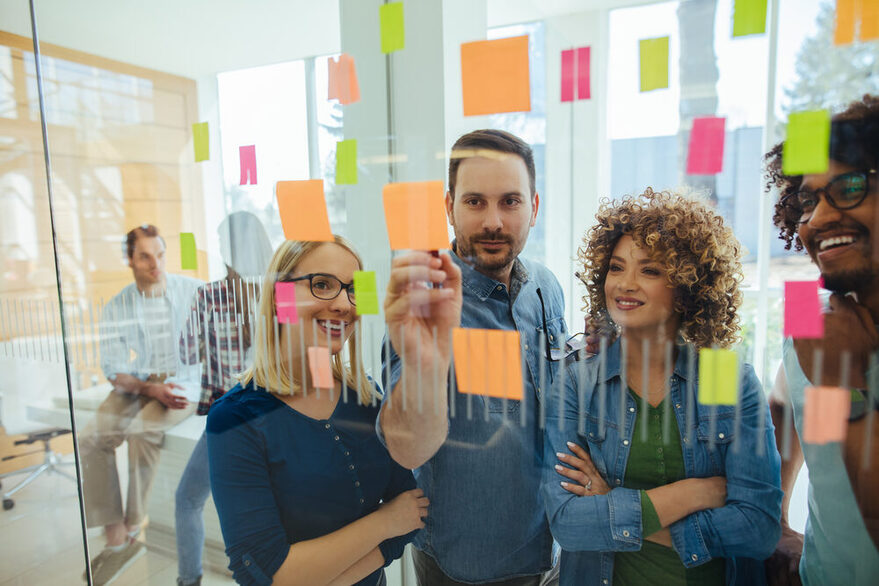 Junge Menschen stehen vor einer Glaswand, auf der Post-It kleben.