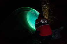 Hintergrund: Ein aufgeschlagenes Zelt bei Nacht. Vordergrund: Eine Person strahlt das Zelt mit einer Taschenlampe an. Die Person trägt eine Jacke mit dem Logo des DRKs auf dem Rücken.