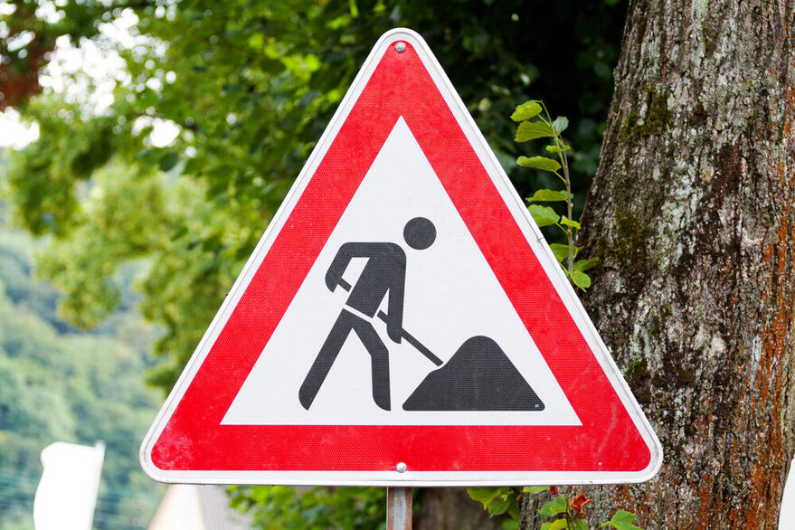 Ein dreeickiges Baustellenschild zeigt ein Strichmännchen, das Erde schaufelt. Das Schild ist rot umrandet. Im Hintergrund ist ein Baum zu erkennen.