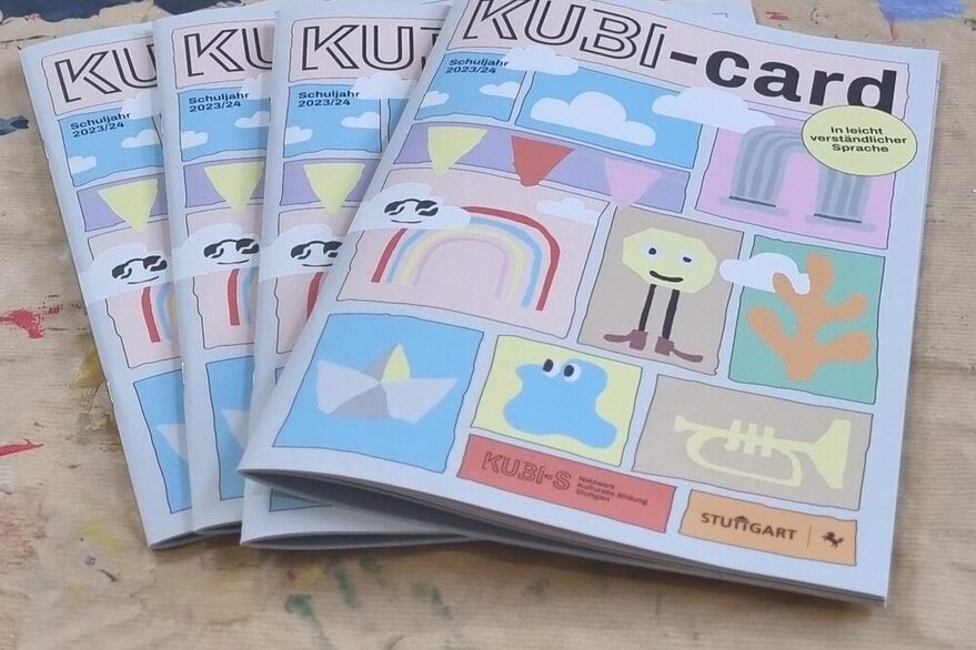 Die KUBI-Card 23-24. Einige Hefte liegen auf einer hölzernen Unterlage, die an einigen Stellen mit Farbkleksen versehen ist. Die Hefte tragen den Schriftzur "KUBI-card" und einige bunte Darstellungen, die von Kindern gemalt wurden.