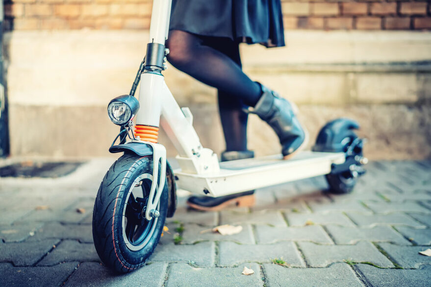 Eine Frau steht mit ihrem Scooter vor einer Hauswand, zu erkennen sind nur ihre Beine.