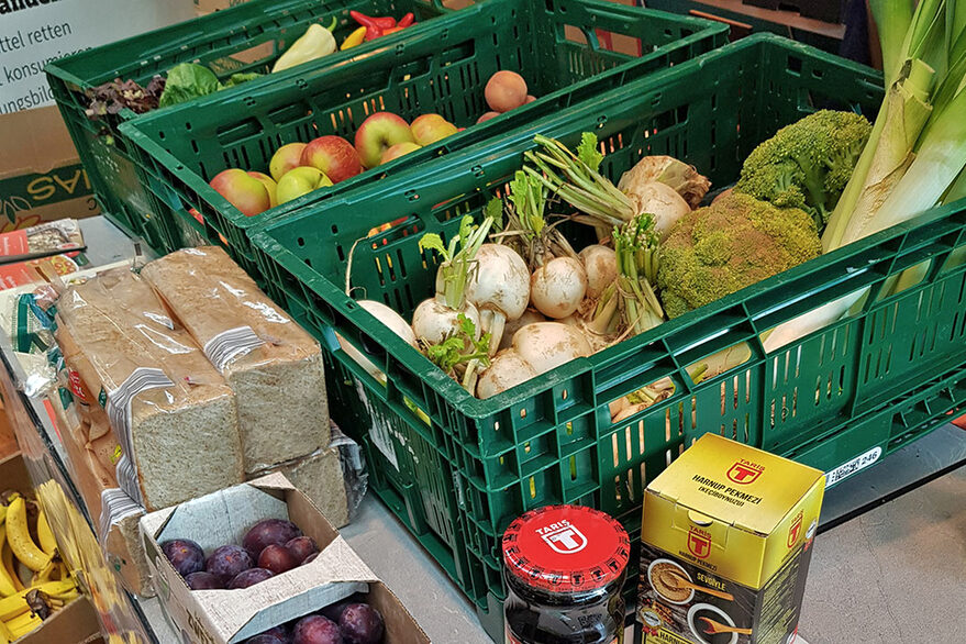 Einige Lebensmittel, wie Äpfel, Rettiche, Pflaumen, Paprika und anderes Gemüse, Brot und Gläser - verstaut in grünen Transportboxen - stehen geordnet im Bild.