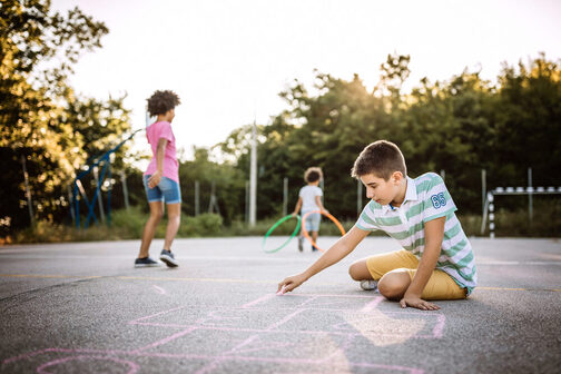 Ein Junge malt mit Kreide auf einem Sportplatz, Kinder spielen im Hintergrund mit bunten Reifen.