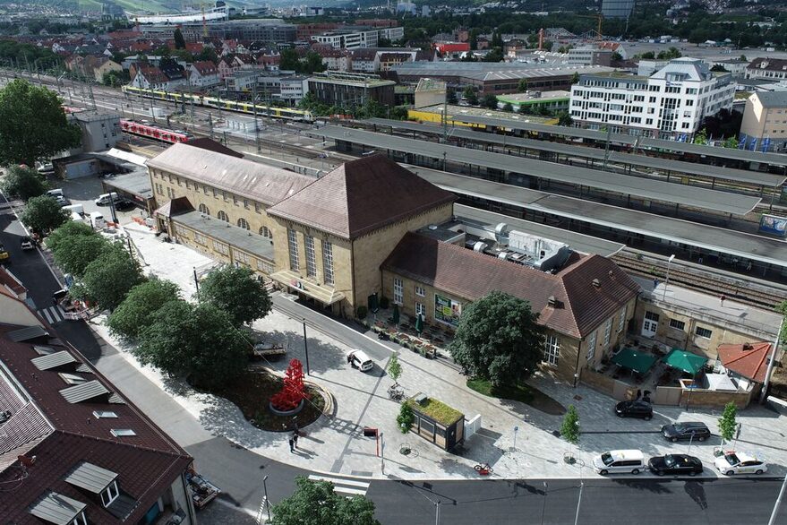 Der neue Bahnhofsvorplatz Bad Cannstatt nach seiner Fertigstellung, fotografiert aus der Vogelperspektive. Der Platz ist hell und mit Bäumen versehen. Einige Menschen und Autos sind ebenfalls zu erkennen, wie auch ein Teil der Gleise mit einem Teil von Bad Cannstatt im Hintergrund.