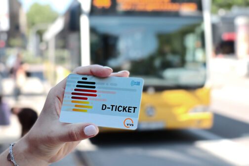 Das Foto zeigt die Hand einer Person vor einem Nahverkehrsbus, die ein Deutschlandticket in der Hand hält.