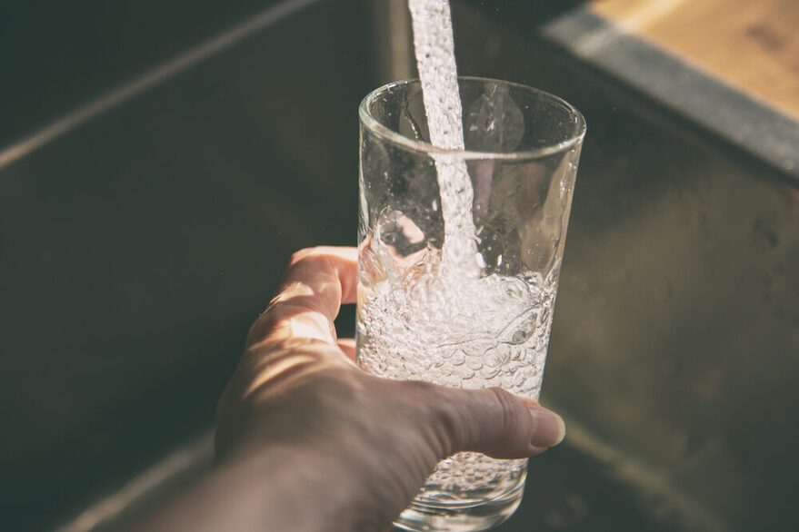 Frisches Wasser aus dem Wasserhahn läuft in ein Glas festgehalten von einer Hand.