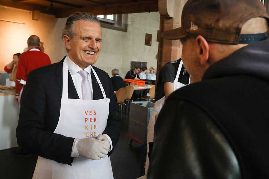 OB Nopper unterhält sich lächelnd mit einem Gast in der Vesperkirche, im Hintergrund verteilen Ehrenamtliche Essen